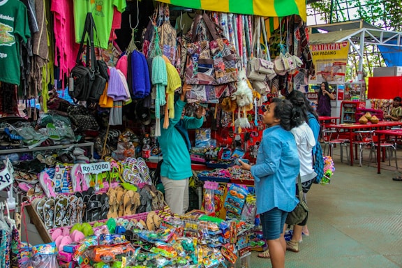 A bustling market scene with various merchandise displayed for sale. There are colorful textiles, bags, and clothing hanging above a wide variety of items including sandals and toys. Several people are browsing and inspecting the items, and the area seems vibrant and full of activity.