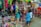 A bustling market scene with various merchandise displayed for sale. There are colorful textiles, bags, and clothing hanging above a wide variety of items including sandals and toys. Several people are browsing and inspecting the items, and the area seems vibrant and full of activity.
