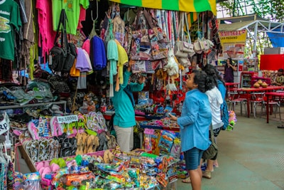 A bustling market scene with various merchandise displayed for sale. There are colorful textiles, bags, and clothing hanging above a wide variety of items including sandals and toys. Several people are browsing and inspecting the items, and the area seems vibrant and full of activity.
