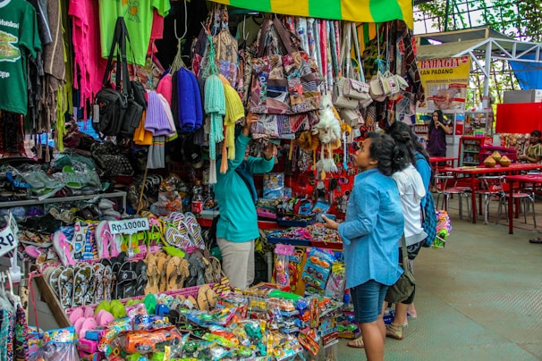 A bustling market scene with various merchandise displayed for sale. There are colorful textiles, bags, and clothing hanging above a wide variety of items including sandals and toys. Several people are browsing and inspecting the items, and the area seems vibrant and full of activity.