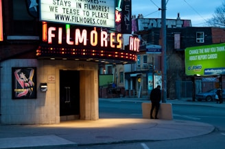 A neon-lit marquee with the name 'Filmore's' and the entrance to an establishment, set against an urban street backdrop. Advertising signs are visible, with a streetscape featuring a pedestrian and a billboard promoting educational services.