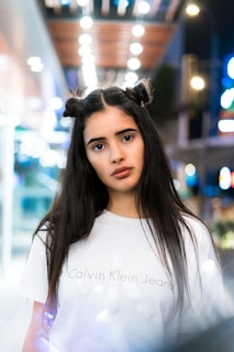 A young person with long dark hair styled in two buns stands in an urban setting at night. The background is blurred with bright bokeh lights, creating a soft and dreamy atmosphere. They are wearing a white Calvin Klein Jeans t-shirt.