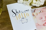 A wooden table surface holds a cute greeting card with the phrase 'She had me at hello' written in playful fonts. Below the text is an illustration of a small, simple bunny shape. To the right, part of a photo shows a sleeping baby wearing a delicate headband. Cream-colored flowers are placed near the top of the image.