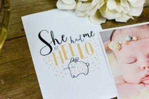 A wooden table surface holds a cute greeting card with the phrase 'She had me at hello' written in playful fonts. Below the text is an illustration of a small, simple bunny shape. To the right, part of a photo shows a sleeping baby wearing a delicate headband. Cream-colored flowers are placed near the top of the image.