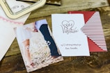 A wooden surface is adorned with a couple of romantic-themed cards. One card shows a photo of a couple holding hands with the word 'Amore' written in cursive. The other card has the phrase 'My Valentine' with a heart graphic and a love note signed 'Love, Samantha'. Additionally, there is a striped red and white envelope partially visible.