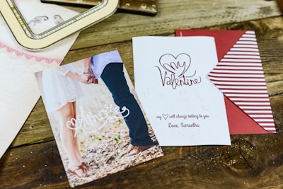 A wooden surface is adorned with a couple of romantic-themed cards. One card shows a photo of a couple holding hands with the word 'Amore' written in cursive. The other card has the phrase 'My Valentine' with a heart graphic and a love note signed 'Love, Samantha'. Additionally, there is a striped red and white envelope partially visible.