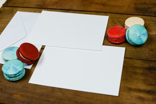 A wooden surface is adorned with several colorful macarons in shades of red, blue, and beige. A partly visible white envelope and two blank white cards accompany them.