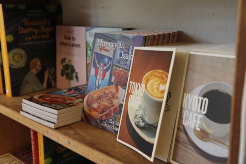 A wooden bookshelf filled with various books, some of which are standing upright while others are stacked horizontally. The book covers display a mix of food-themed and instructional titles, such as 'Tokyo Cafe', 'Kyoto Cafe', and 'How to raise a plant'. The shelf carries a casual, cozy vibe, suggesting a focus on lifestyle and culinary topics.