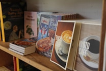 A wooden bookshelf filled with various books, some of which are standing upright while others are stacked horizontally. The book covers display a mix of food-themed and instructional titles, such as 'Tokyo Cafe', 'Kyoto Cafe', and 'How to raise a plant'. The shelf carries a casual, cozy vibe, suggesting a focus on lifestyle and culinary topics.