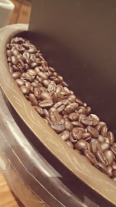 A close-up view of glossy, roasted coffee beans piled in a decorative bowl, featuring intricate patterns on its rim. The image captures the rich textures and dark hues of the beans with a warm, inviting ambiance.