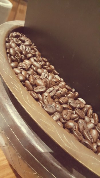 A close-up view of glossy, roasted coffee beans piled in a decorative bowl, featuring intricate patterns on its rim. The image captures the rich textures and dark hues of the beans with a warm, inviting ambiance.