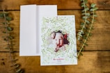A greeting card with a photo of a baby framed by a decorative wreath of green leaves and small red bows. The card is placed on a wooden surface with a white envelope next to it and two sprigs of greenery on either side.