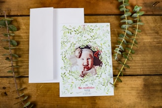 A greeting card with a photo of a baby framed by a decorative wreath of green leaves and small red bows. The card is placed on a wooden surface with a white envelope next to it and two sprigs of greenery on either side.