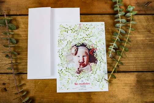 A greeting card with a photo of a baby framed by a decorative wreath of green leaves and small red bows. The card is placed on a wooden surface with a white envelope next to it and two sprigs of greenery on either side.