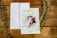 A greeting card with a photo of a baby framed by a decorative wreath of green leaves and small red bows. The card is placed on a wooden surface with a white envelope next to it and two sprigs of greenery on either side.