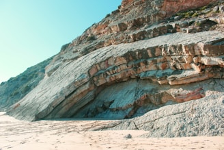 A coastal scene featuring a large rock formation with distinct layers and textures. The rock is set against a clear blue sky, and the foreground includes sandy terrain with some visible footprints.