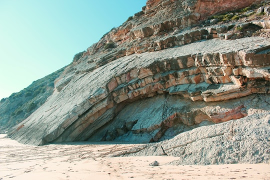A coastal scene featuring a large rock formation with distinct layers and textures. The rock is set against a clear blue sky, and the foreground includes sandy terrain with some visible footprints.