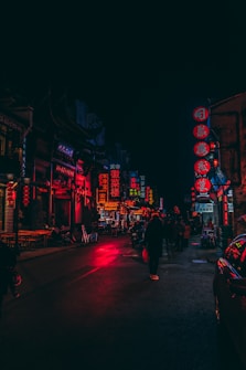 A vibrant urban street scene at night, illuminated by numerous neon signs in shades of red and blue. People walk along the dimly lit street, with vehicles parked on the sides. The atmosphere is bustling yet moody, characteristic of a lively nightlife environment.