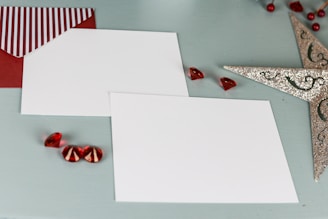 Two blank white cards are placed on a light blue background. Beside them is a red envelope with a striped flap. Decorative red gems and a sparkling silver star-shaped ornament are arranged around the cards, giving a festive appearance.