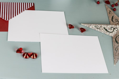 Two blank white cards are placed on a light blue background. Beside them is a red envelope with a striped flap. Decorative red gems and a sparkling silver star-shaped ornament are arranged around the cards, giving a festive appearance.