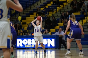 A group of female basketball players are on a court, with one player in a white uniform holding the basketball above her head, preparing to pass or shoot. The gymnasium's bleachers are filled with spectators and yellow-blue decor. A scoreboard with the word 'ROBINSON' is visible in the background.