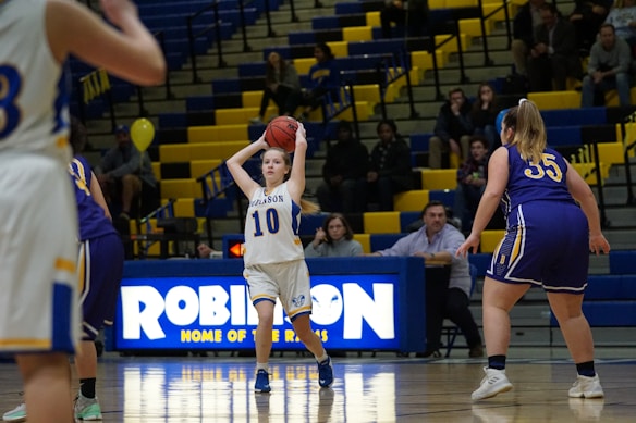 A group of female basketball players are on a court, with one player in a white uniform holding the basketball above her head, preparing to pass or shoot. The gymnasium's bleachers are filled with spectators and yellow-blue decor. A scoreboard with the word 'ROBINSON' is visible in the background.