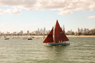 A red sailboat is sailing across the water with several other boats in the background. The skyline of a city with tall buildings is visible under a partly cloudy sky.