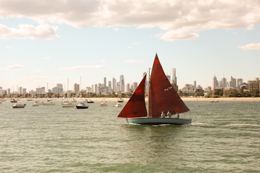 A red sailboat is sailing across the water with several other boats in the background. The skyline of a city with tall buildings is visible under a partly cloudy sky.