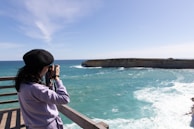 A person wearing a black beret and purple sweater is using a camera to photograph a rocky coastline. The water appears turquoise under the bright sky. The person is positioned on a wooden deck, suggesting an observation point or lookout.