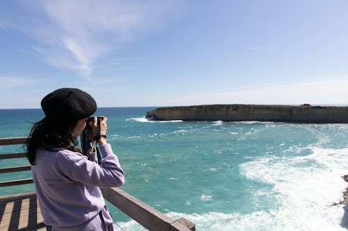 A person wearing a black beret and purple sweater is using a camera to photograph a rocky coastline. The water appears turquoise under the bright sky. The person is positioned on a wooden deck, suggesting an observation point or lookout.