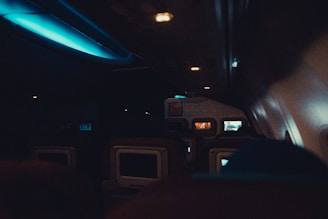 The interior of an airplane cabin with several rows of seats visible. Ambient lighting creates a dim atmosphere, with a blue glow from overhead lights. Seatback screens suggest passengers may be flying in an economy or business class. Some screens display content, while others are turned off.
