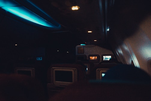 The interior of an airplane cabin with several rows of seats visible. Ambient lighting creates a dim atmosphere, with a blue glow from overhead lights. Seatback screens suggest passengers may be flying in an economy or business class. Some screens display content, while others are turned off.