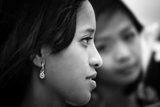 A black and white profile of a young woman with long hair elegantly styled, wearing a decorative earring. Another person is blurred in the background, creating depth and focus on the main subject.