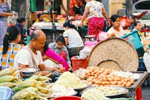 A bustling outdoor market scene with several people engaged in various activities. Fresh produce, including corn, eggs, bamboo shoots, and other vegetables, is displayed prominently on tables. The scene is vibrant with people wearing colorful clothing and some sitting or standing behind their goods. The background suggests a typical market atmosphere with more goods and people.