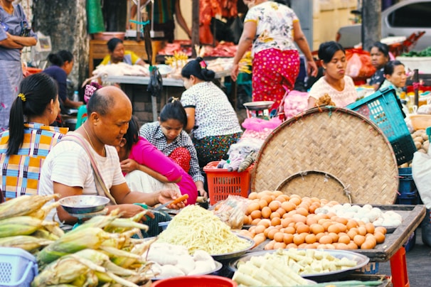 A bustling outdoor market scene with several people engaged in various activities. Fresh produce, including corn, eggs, bamboo shoots, and other vegetables, is displayed prominently on tables. The scene is vibrant with people wearing colorful clothing and some sitting or standing behind their goods. The background suggests a typical market atmosphere with more goods and people.