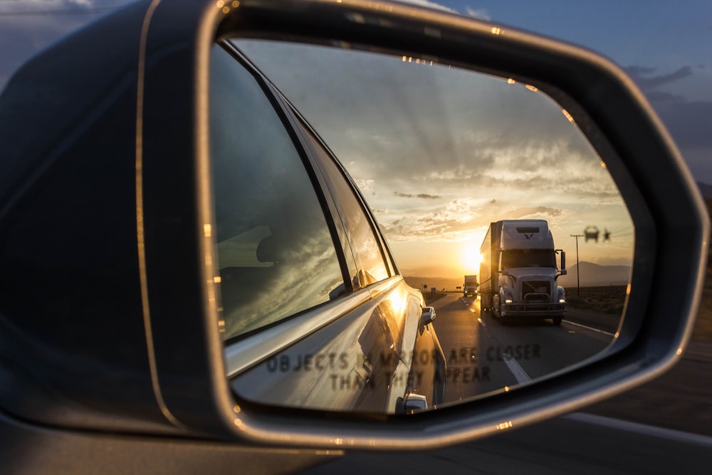 reflection of white freight truck on grey car wing mirror during orange sunset