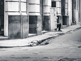 A black and white image depicts a street scene with a solitary figure sitting against a wall, holding a mobile phone. The street is empty, and the buildings have a worn, aged appearance. The figure is wearing casual clothing and sneakers, and the environment conveys a sense of urban solitude.