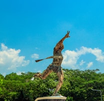 A statue of a runner breaking through a finish line, with arms raised in a triumphant pose. The sculpture is set against a vibrant blue sky with scattered clouds and lush green foliage below.