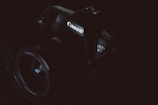 A Canon camera is partially visible in low lighting. The focus is on the branding and the model number EOS 6D, which is subtly illuminated. The dark and moody composition highlights the contours and details of the camera body.