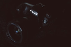 A Canon camera is partially visible in low lighting. The focus is on the branding and the model number EOS 6D, which is subtly illuminated. The dark and moody composition highlights the contours and details of the camera body.