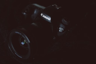 A Canon camera is partially visible in low lighting. The focus is on the branding and the model number EOS 6D, which is subtly illuminated. The dark and moody composition highlights the contours and details of the camera body.