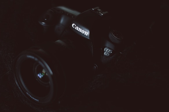 A Canon camera is partially visible in low lighting. The focus is on the branding and the model number EOS 6D, which is subtly illuminated. The dark and moody composition highlights the contours and details of the camera body.