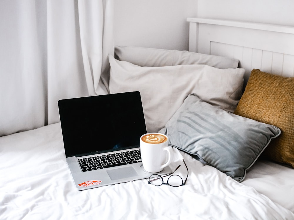 寝室の白いベッドの上の灰色の枕の近くにMacBook Proでコーヒーを飲んだ白いセラミックのマグカップ