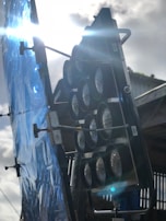 A large lighting fixture with multiple circular elements is positioned outdoors. It is mounted on a stand beside a reflective blue sheet that seems to be used for diffusing light. The sun is shining brightly, casting reflections and lens flares across the image, revealing an industrial setting with a metallic structure in the background.