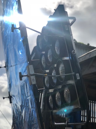 A large lighting fixture with multiple circular elements is positioned outdoors. It is mounted on a stand beside a reflective blue sheet that seems to be used for diffusing light. The sun is shining brightly, casting reflections and lens flares across the image, revealing an industrial setting with a metallic structure in the background.