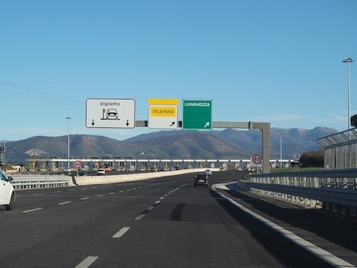 A highway scene features a toll booth area with multiple lanes, some designated for ticket collection and others for Telepass electronic payment. Overhead signs display directions, including one for Lunghezza. Traffic signs indicate a speed limit of 60 km/h. The setting is surrounded by mountains under a clear blue sky.