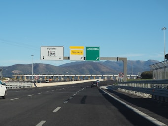A highway scene features a toll booth area with multiple lanes, some designated for ticket collection and others for Telepass electronic payment. Overhead signs display directions, including one for Lunghezza. Traffic signs indicate a speed limit of 60 km/h. The setting is surrounded by mountains under a clear blue sky.