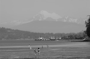 A black and white image featuring a beach with several people walking on the sand. In the background, there is a mountain range with snowy peaks, and a line of boats docked near the shoreline. The scene is complemented by a forested area on the right side.