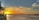 A scenic sunset over a calm body of water with a vibrant orange and yellow sky. Chinese fishing nets are visible on the left, along with a person standing on the shore. Distant buildings and silhouettes of trees line the horizon.