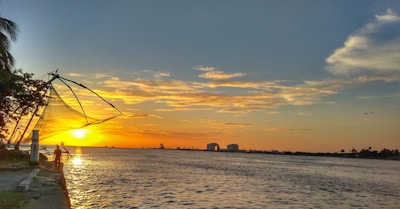 A scenic sunset over a calm body of water with a vibrant orange and yellow sky. Chinese fishing nets are visible on the left, along with a person standing on the shore. Distant buildings and silhouettes of trees line the horizon.
